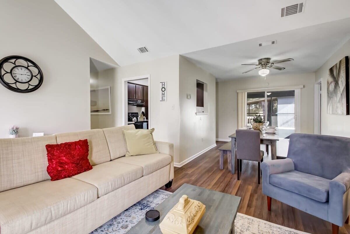 Un salon avec un canapé beige, un coussin rouge, une table basse et un fauteuil bleu. On y trouve également un coin repas avec une table et des chaises, et une cuisine à l’arrière-plan.