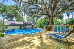 Una terraza de madera con piscina, sillones de descanso y una casa con cobertizo.