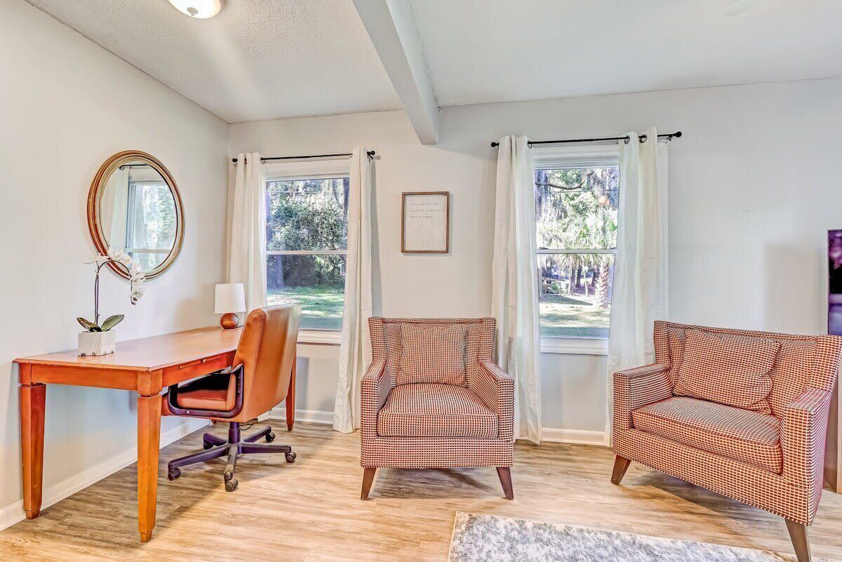 Una habitación con un escritorio de madera, dos sillones estampados, un espejo y un cuadro en la pared.