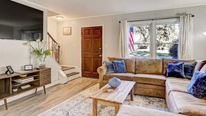 Un salon avec un canapé, une table basse et un téléviseur. Il y a une porte et une fenêtre, et on peut voir un drapeau américain.