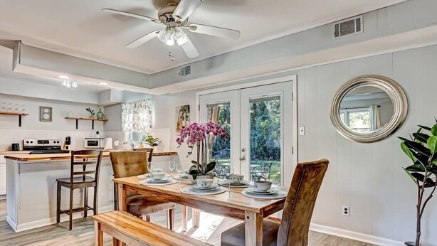 Un coin repas avec une table en bois dressée pour quatre personnes, un ventilateur de plafond et une vue sur l’extérieur grâce à de grandes fenêtres.