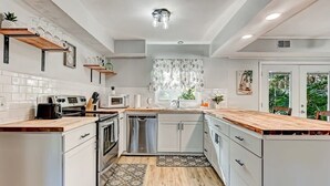 Une cuisine moderne avec des meubles blancs, un plan de travail en bois, des appareils en acier inoxydable et une fenêtre agrémentée d’un rideau à motifs floraux.