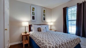 Une chambre à coucher avec un lit, deux tables de chevet avec des lampes, une fenêtre avec des rideaux et deux tableaux encadrés accrochés au mur.