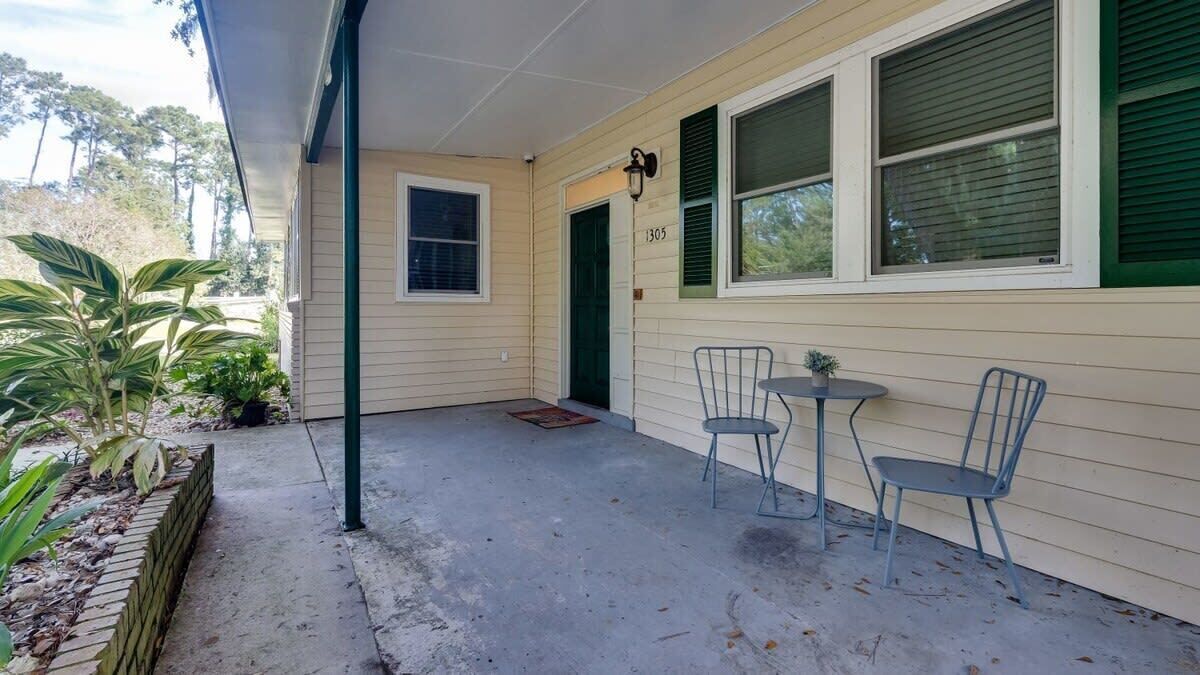 A covered porch with two metal chairs and a small table, a door with the number 1305, and a brick planter with plants.