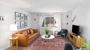 Una sala de estar con un sofá de cuero marrón, una mesa de centro redonda, un sillón verde, un televisor sobre un mueble de madera y una alfombra estampada.