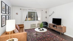Una sala de estar moderna con un sofá de cuero marrón, una mesa redonda, un televisor sobre un mueble de madera y un ventanal con cortinas blancas.