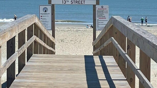 Une promenade en bois mène à une plage où un panneau indique « 13e rue » et où sont affichées les règles à respecter sur la plage.