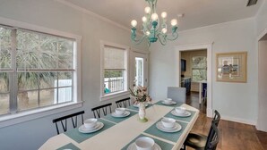 A dining room with a long table set for four, a chandelier, and a view of the outdoors through large windows. - Walk to Beach! Flamingle in Fuchsia, Whole House (Tybee Island)