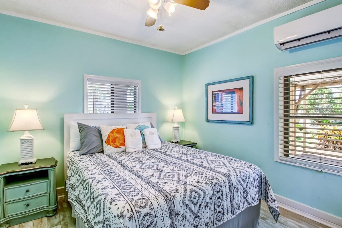 A bedroom with a bed, two bedside tables with lamps, a ceiling fan, a window with blinds, and an air conditioning unit.