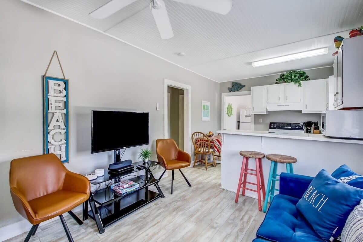 Ein modernes Wohnzimmer mit einem Flachbildfernseher, Ledersesseln, einem Couchtisch und einem blauen Sofa mit Kissen mit der Aufschrift 'BEACH'.