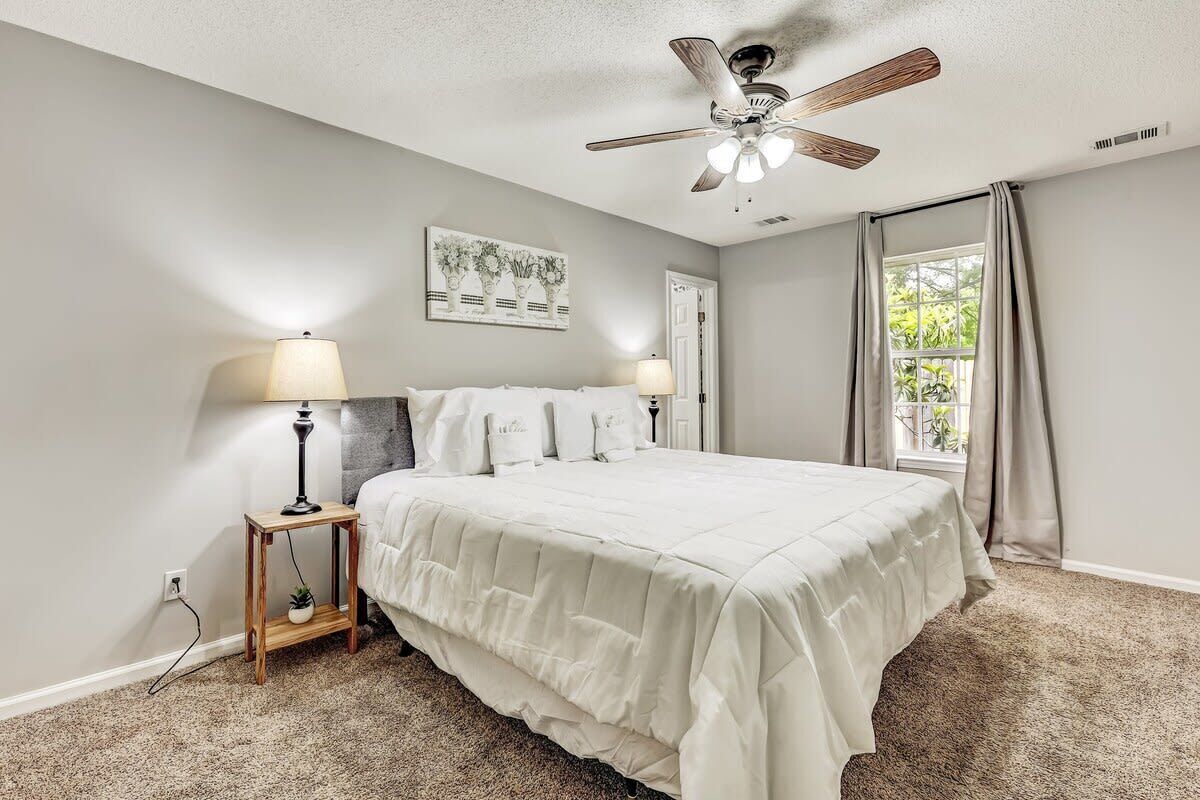 A bedroom with a bed, two bedside lamps, a ceiling fan, a window with curtains, and a wall-mounted artwork.