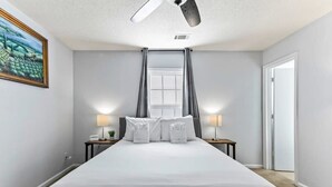 Una habitación de hotel con una cama grande, dos mesitas de noche con lámparas, un ventilador de techo, una ventana con cortinas y una puerta.