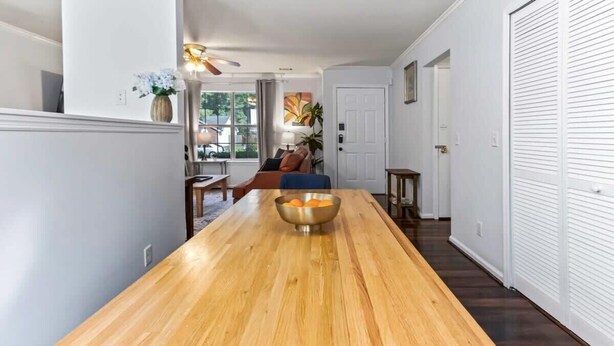 Una larga mesa de comedor de madera con un bol de naranjas en el centro, un ventilador de techo y vistas a una sala de estar con un sofá y una planta.