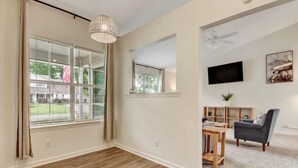 A spacious living room with a large window, a ceiling fan, a flat-screen TV, and a grey sofa.