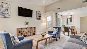A spacious living room with a sofa, armchairs, a coffee table, and a TV mounted on the wall. There is a dining area with a table and chairs visible in the background.