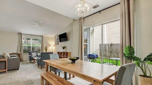 A spacious living room with a wooden dining table, comfortable seating, a ceiling fan, a TV mounted on the wall, and large windows with curtains.