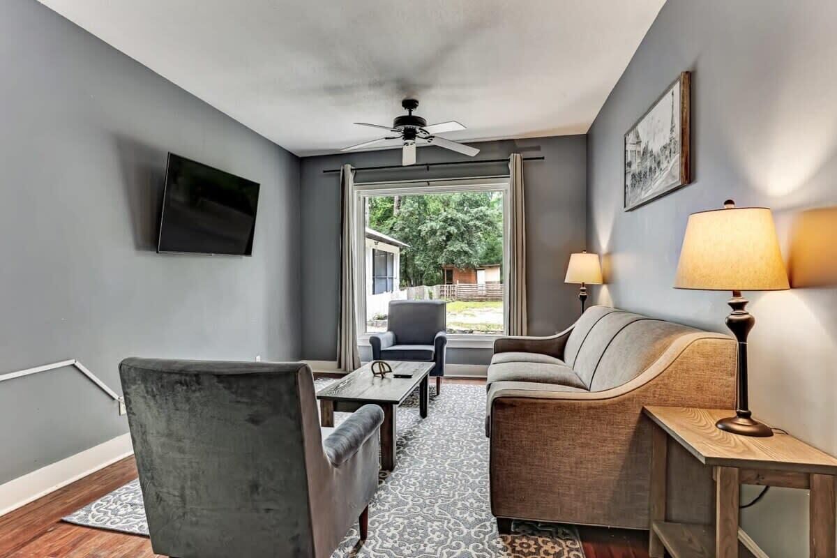 A living room with a grey sofa, a grey armchair, a wooden coffee table, a TV mounted on the wall, and a ceiling fan.