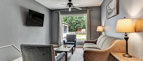 A living room with a grey sofa, a grey armchair, a wooden coffee table, a TV mounted on the wall, and a ceiling fan.