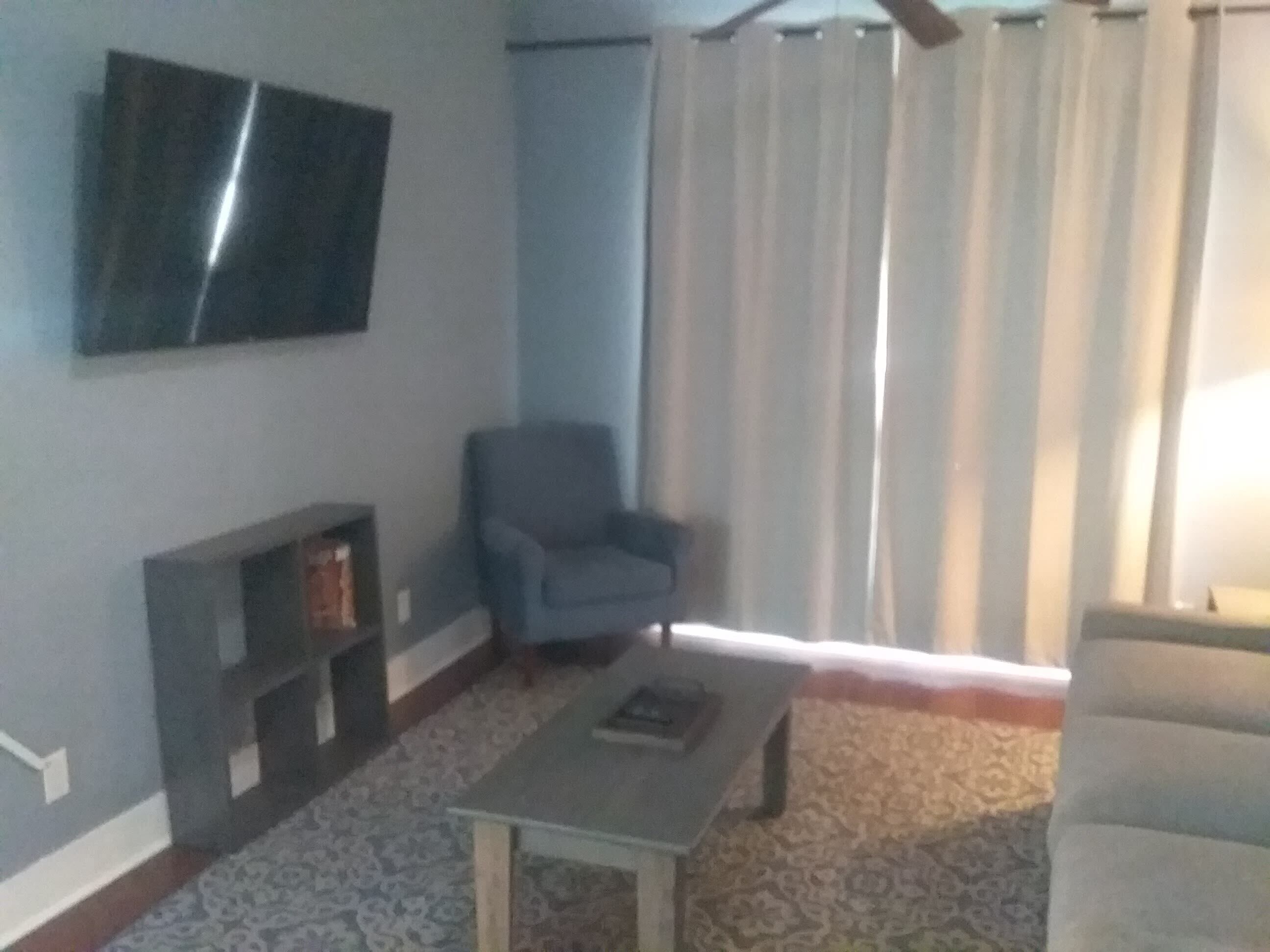 A living room with a flat-screen TV mounted on the wall, a blue armchair, a patterned carpet, and a small table.