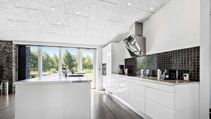A modern kitchen with a central island, white cabinetry, a black backsplash, and a view of greenery outside. - Awesome home in Glesborg with sauna (Glesborg)
