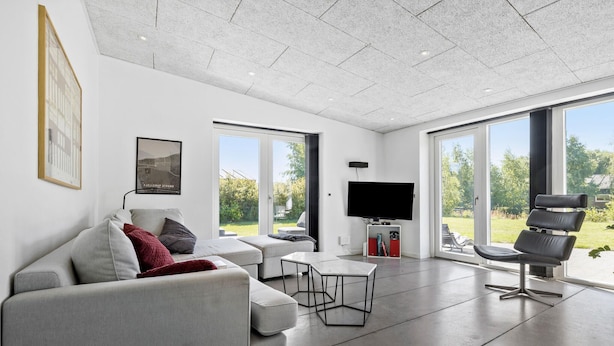 A modern living room with a sofa, a coffee table, and a TV. There are large windows and a view of the outdoors. - Awesome home in Glesborg with sauna (Glesborg)
