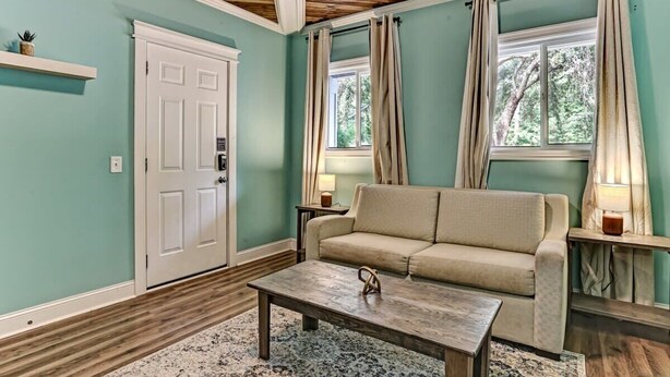 Ein Wohnzimmer mit türkisfarbenen Wänden, einer Holzbalkendecke, einem beigen Sofa, einem Couchtisch und zwei mit Vorhängen versehenen Fenstern.