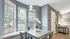 A dining area with a table and chairs, a potted plant, and large windows with blinds.