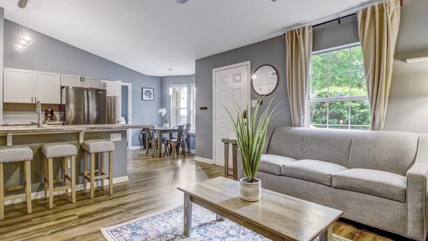 A modern kitchen with stainless steel appliances, a dining area with a table and chairs, a living room with a sofa and a coffee table, and a large window with curtains.