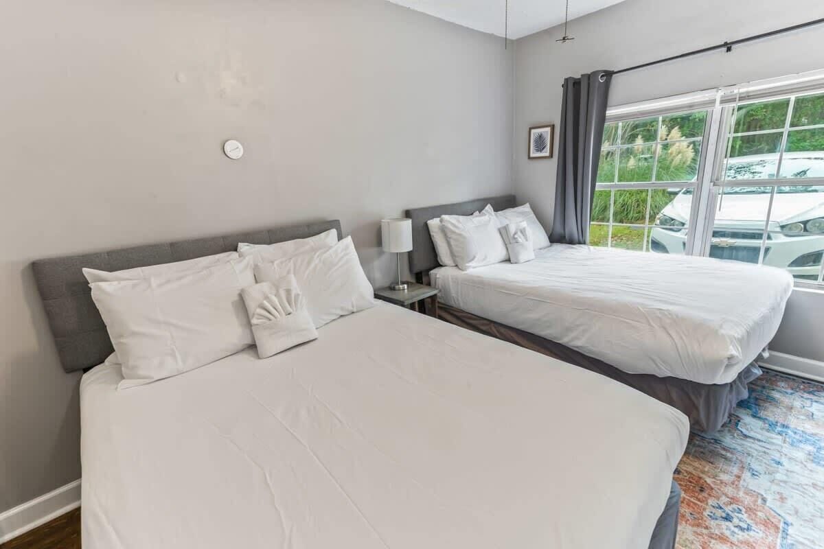 A hotel room with two beds, a large window with a view of a car and greenery, a bedside table with a lamp, and a wall clock.