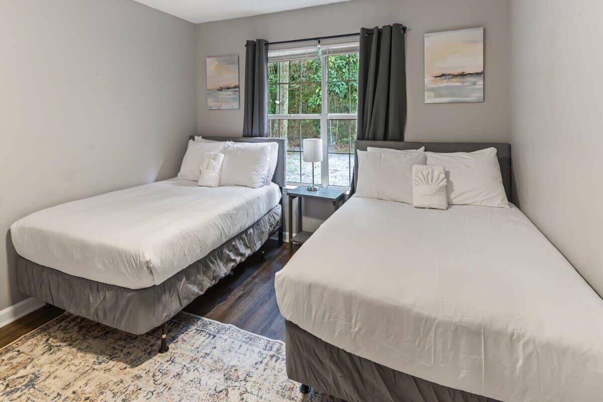 A hotel room with two single beds, a small table, a window with curtains, and a view of greenery outside.