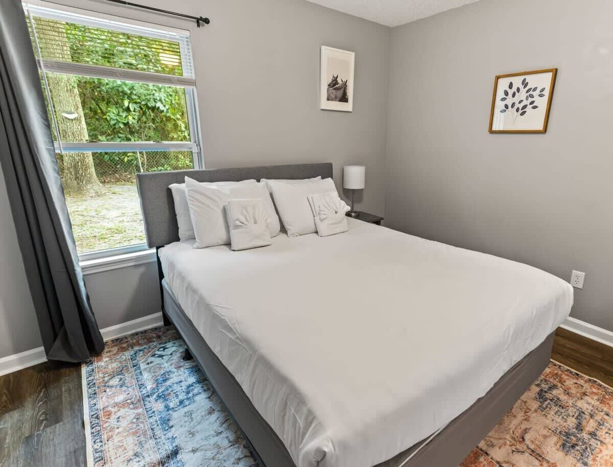 A bedroom with a large bed, a window with curtains, a bedside lamp, and a framed picture on the wall.