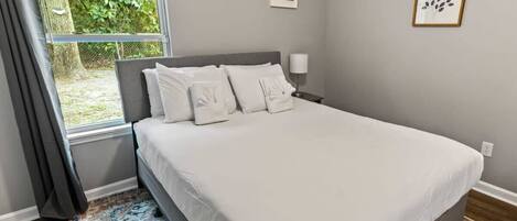 A bedroom with a large bed, a window with curtains, a bedside lamp, and a framed picture on the wall.