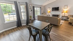 A spacious living room with a wooden dining table and chairs, a fireplace, and a television mounted on the wall. - Fenced Yard & BBQ Grill Near Dining, Parks & Shops (Savannah)