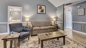 Ein Wohnzimmer mit grauem Sofa, blauem Sessel und Couchtisch aus Holz. Zwei Stehlampen und ein gerahmtes Bild sind an der Wand zu sehen.