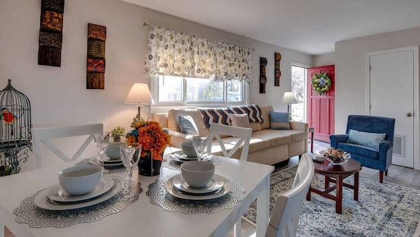 A dining area with a table set for two, a sofa with cushions, a blue armchair, and a red door.
