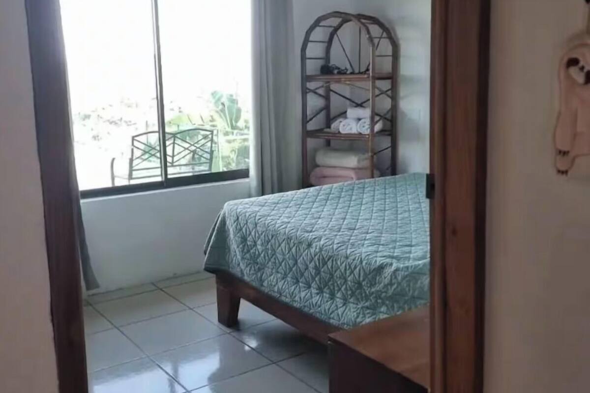 A bedroom with a bed, a window, a shelf, and a view of the outdoors.