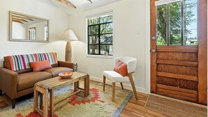 Sala de estar com um sofá de couro marrom, uma poltrona branca, uma mesa de centro de madeira e um tapete colorido. Há uma porta de madeira com painéis de vidro e uma janela com vista para árvores.