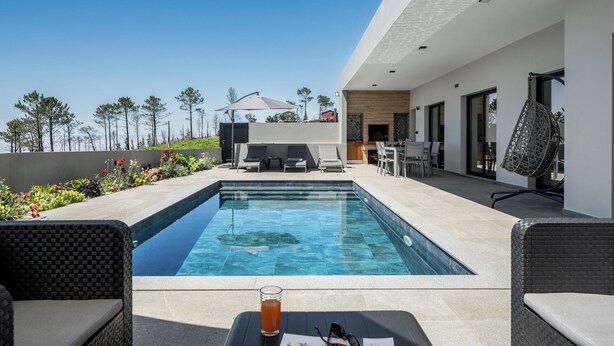 Un área de piscina moderna al aire libre con una piscina azul cristalina, sillas de descanso y una mesa de comedor con una bebida.