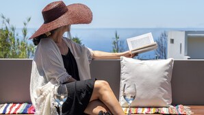 Una persona sentada en un balcón, leyendo un libro, con una copa de vino y un cojín decorativo.