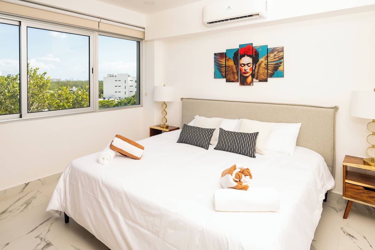 A hotel room with a large bed, a view of greenery and buildings through the window, a wall with a painting, and a ceiling fan.