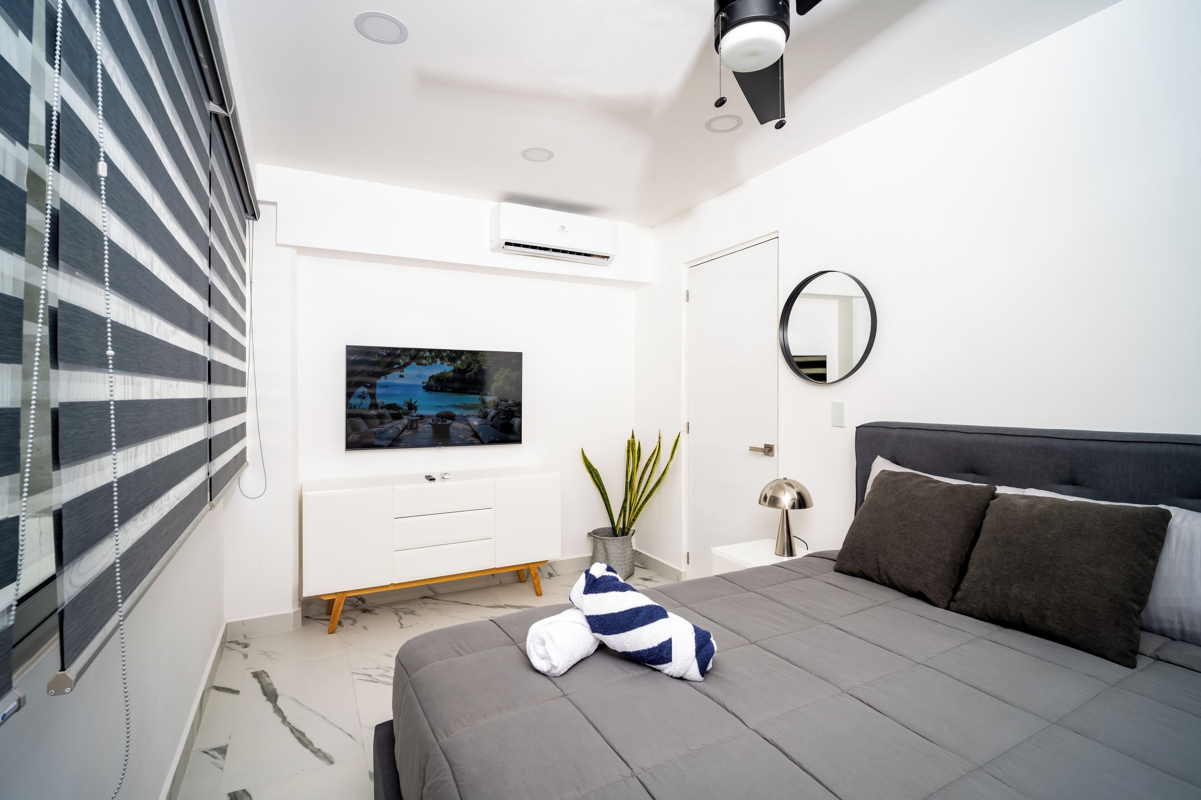 A modern bedroom with a grey bed, a TV mounted on the wall, a potted plant, and a bedside table with a lamp.