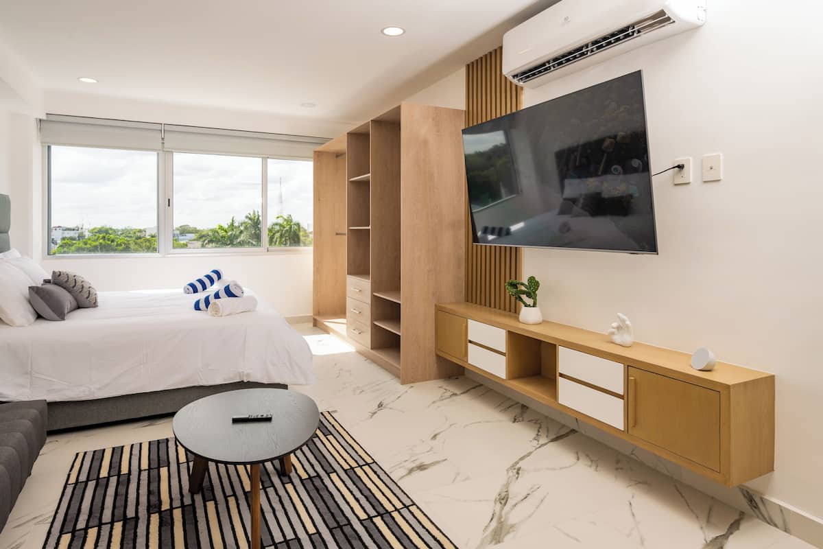 A modern bedroom with a bed, a TV mounted on the wall, a built-in wooden cabinet, and a large window providing a view of greenery outside.