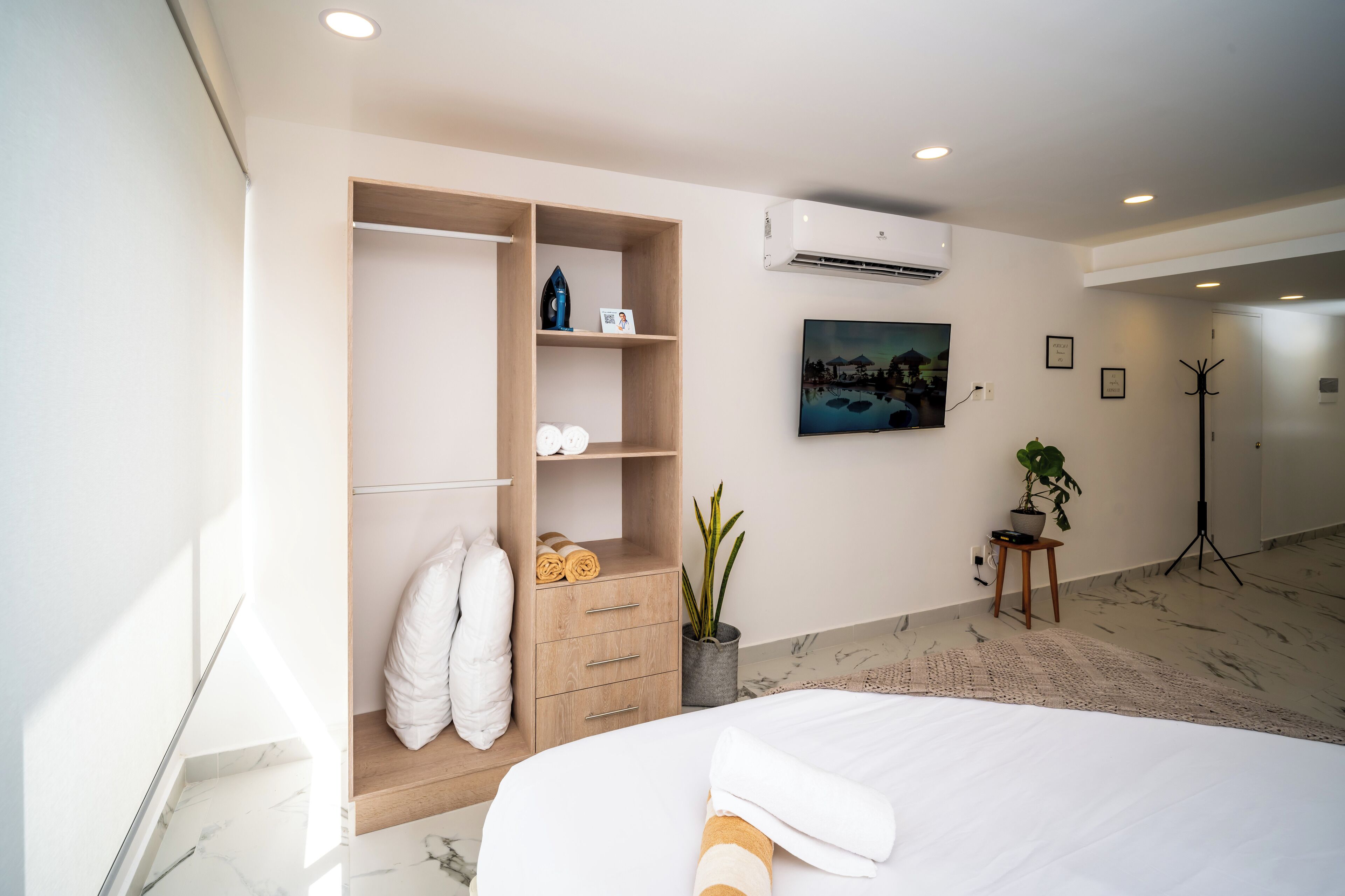 A modern hotel room with a bed, a wooden wardrobe, a potted plant, a painting on the wall, and an air conditioning unit.