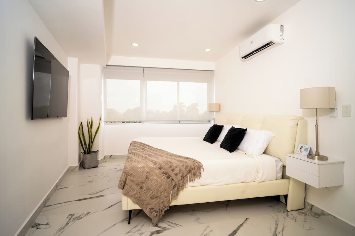 A modern bedroom with a large bed, a TV mounted on the wall, a window with a view, and an air conditioning unit.