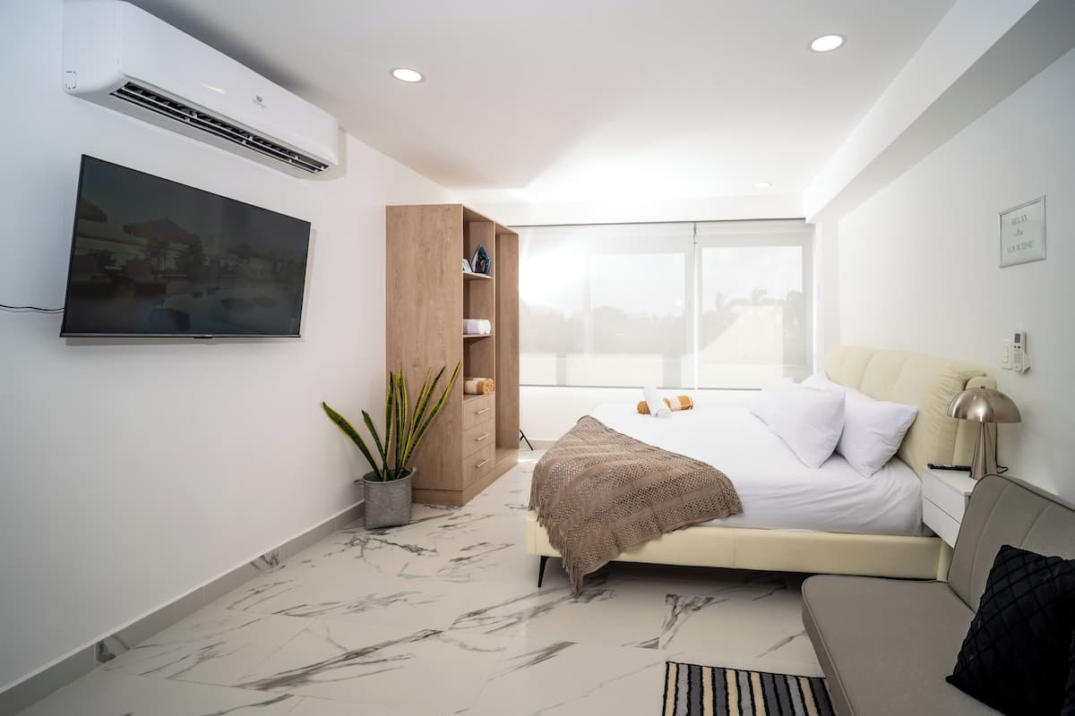A modern bedroom with a large bed, a TV mounted on the wall, a wooden cabinet, and a large window offering a city view.