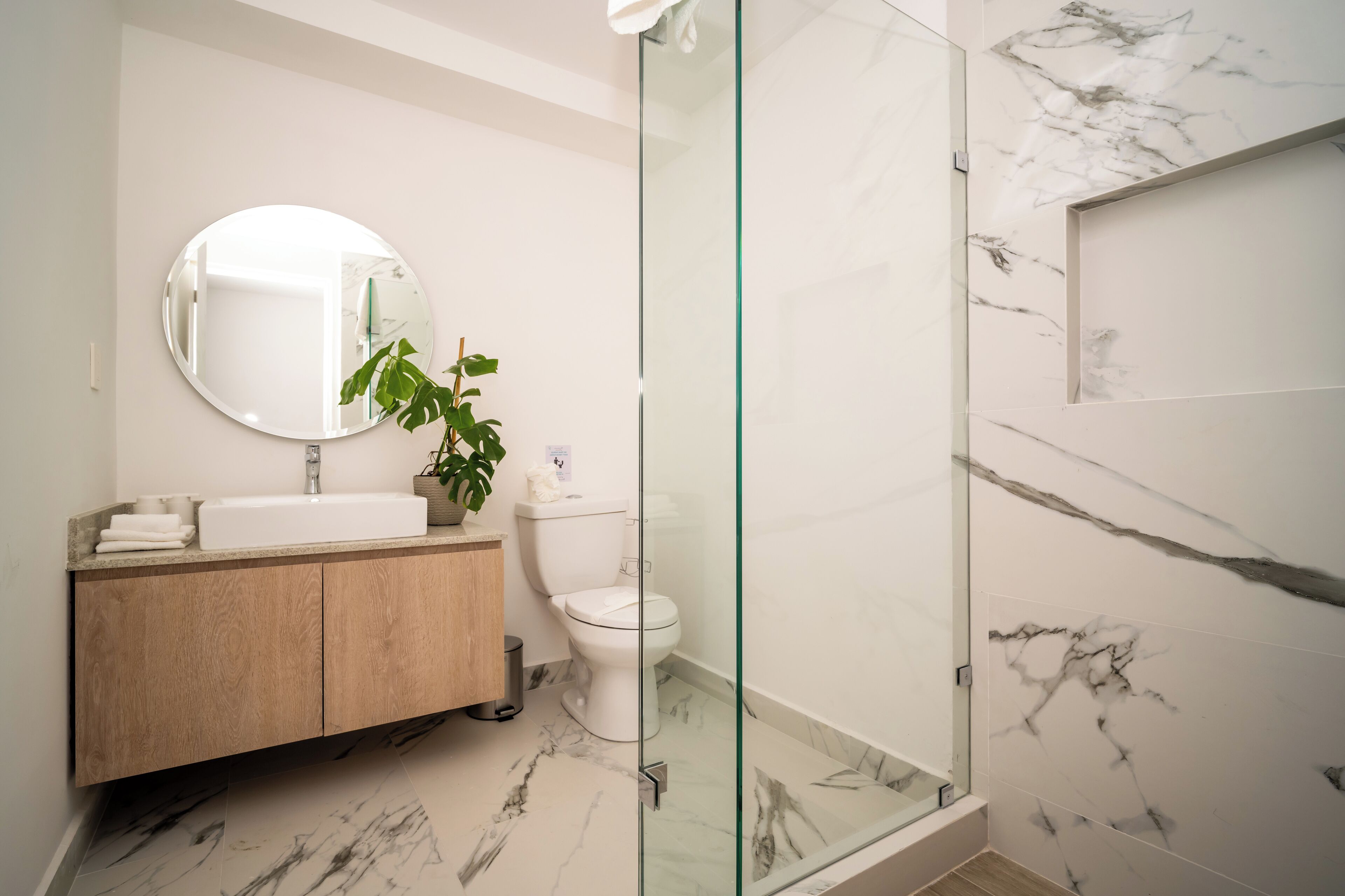 A modern bathroom with a marble floor, a wooden vanity, a round mirror, a toilet, and a glass shower enclosure.