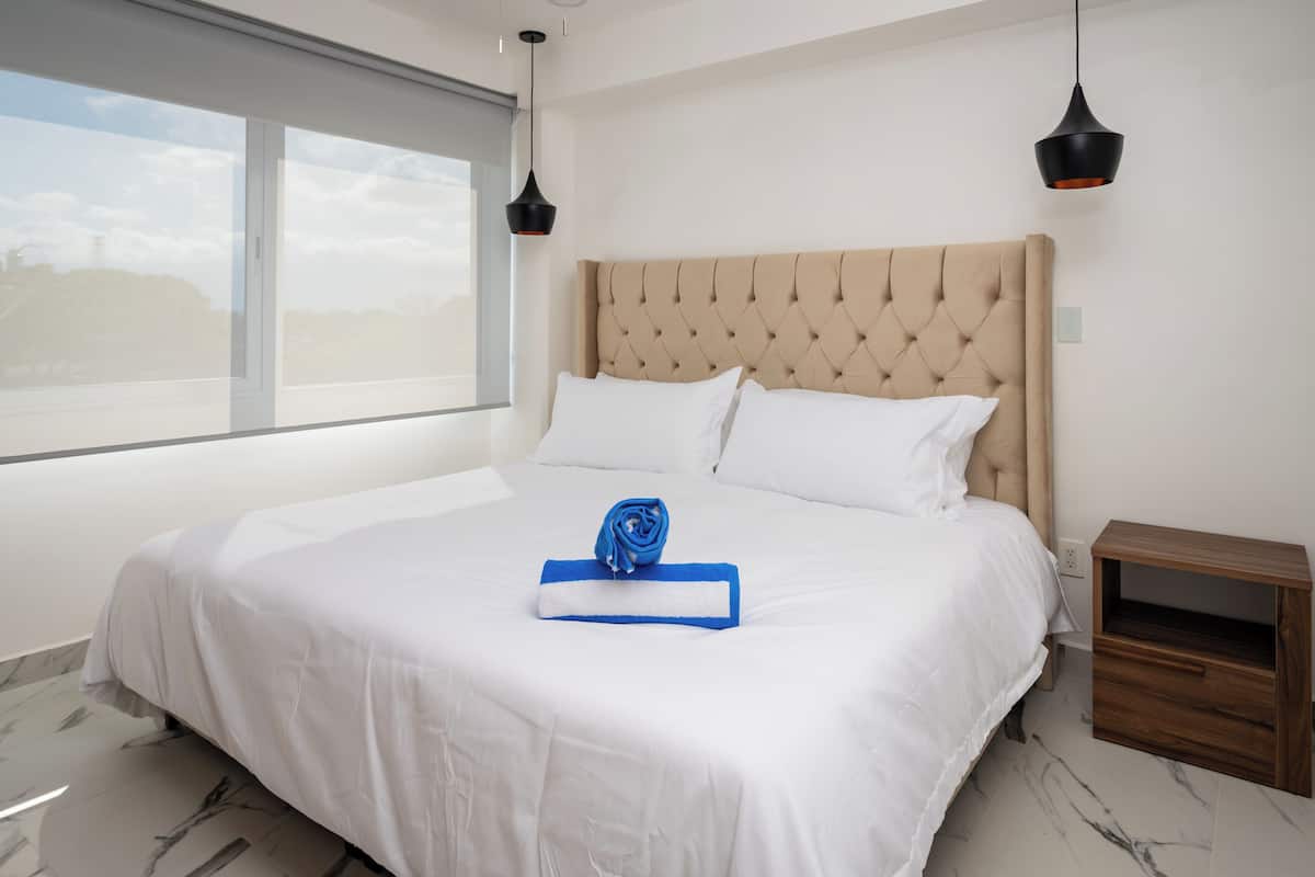 A neatly made bed with a blue towel and a rolled-up towel, a wooden nightstand, and a large window with a view of the outdoors.