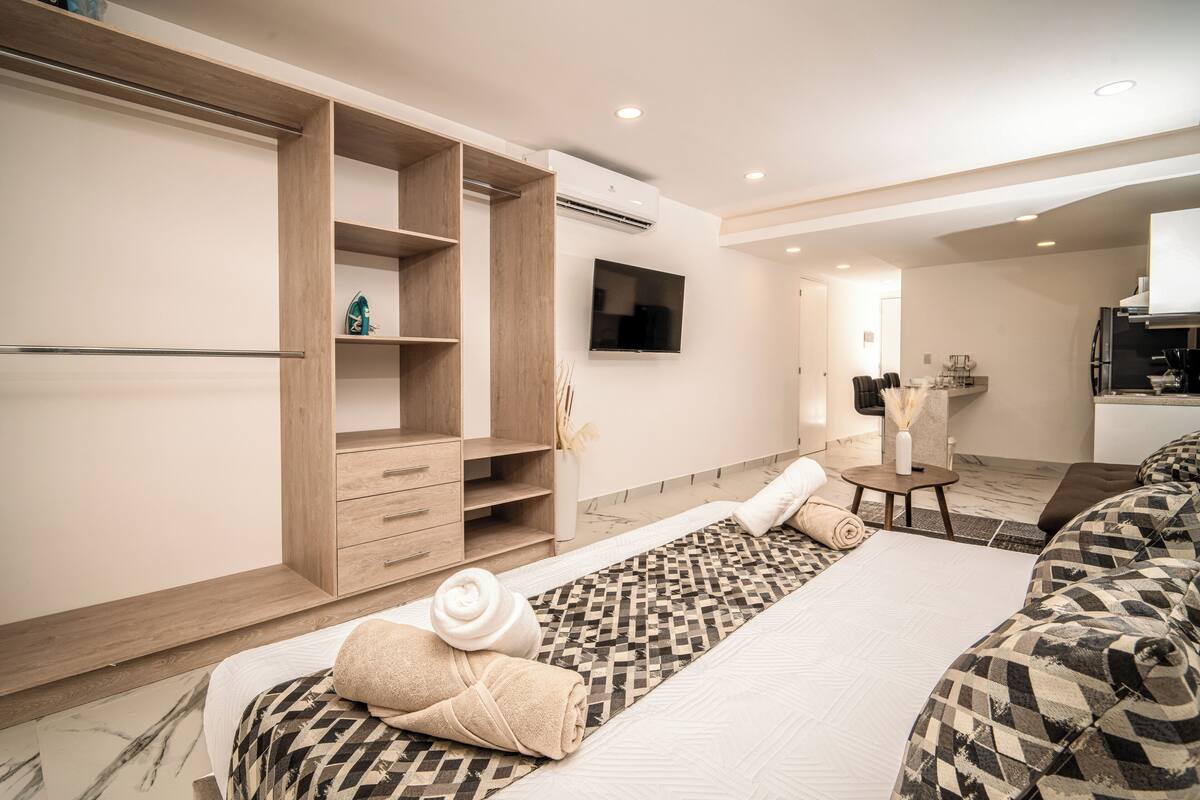 A modern hotel room with a bed, a TV, a wardrobe, and a small kitchenette.