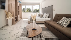 A modern living room with a sofa, a coffee table, a TV, and a large window with a view of palm trees. - Barcelona Condos Cancun (Cancún)