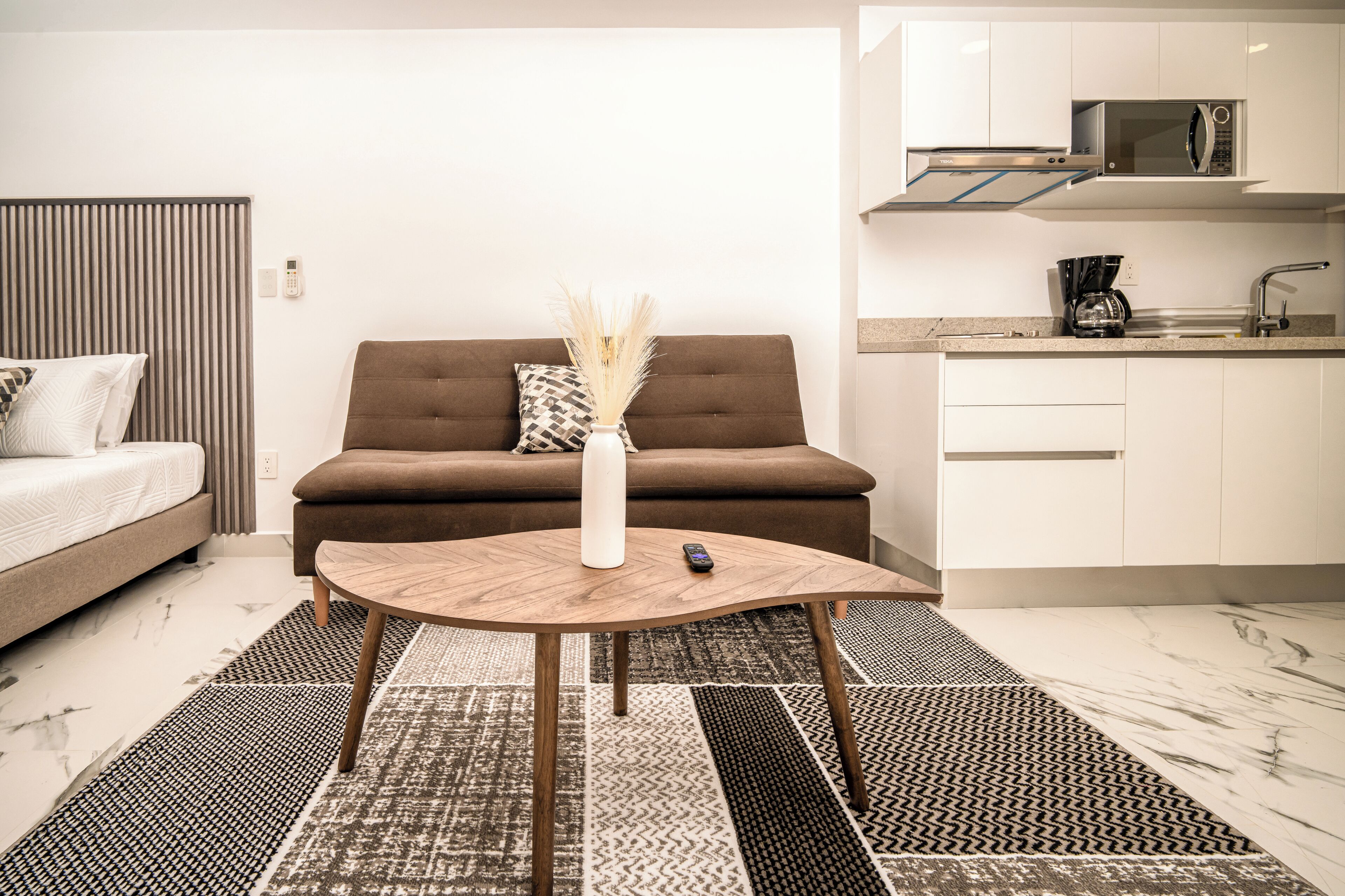 A modern living room with a sofa, a coffee table, a vase with dried plants, and a kitchen area with a microwave and coffee maker.